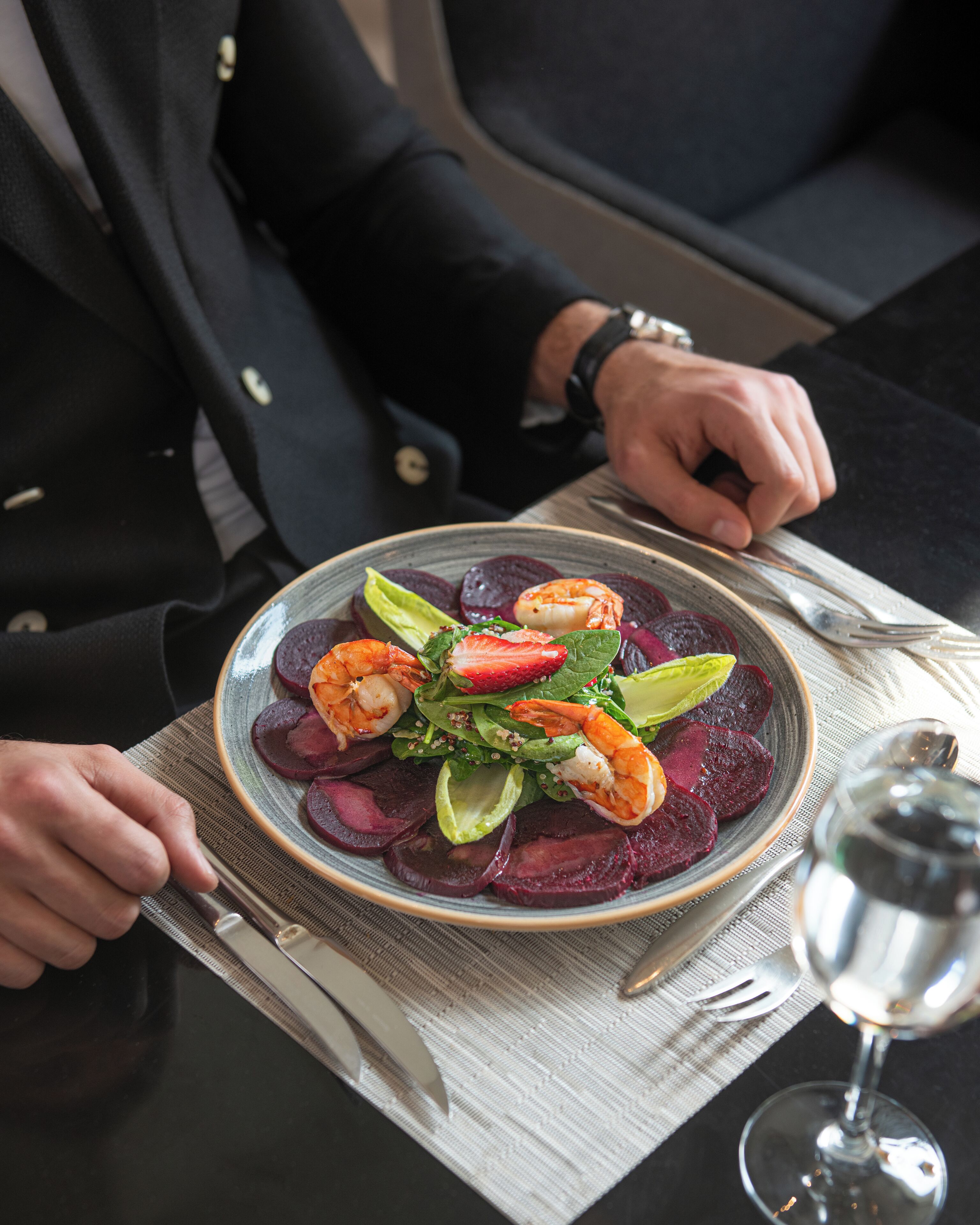 A plate of food with shrimp, beets, and greens, a knife, and a glass of water on a table.