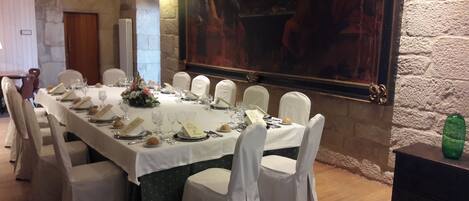 A long dining table set for a formal meal with white tablecloths, chairs, and a large painting on the wall.