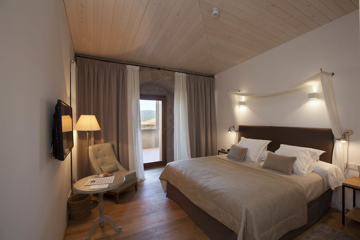 Een hotelkamer met een groot bed, een stoel, een klein tafeltje en uitzicht door een deur.