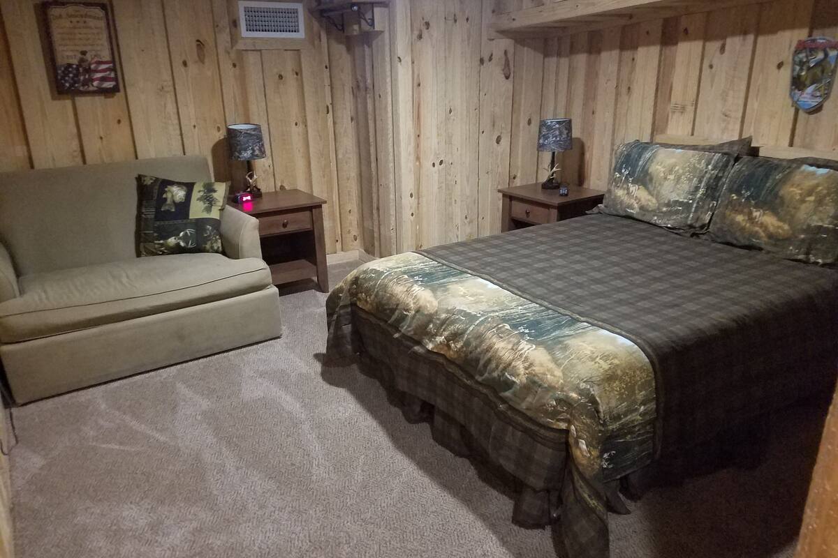Ein Zimmer mit Holzwänden, einem Bett mit Tarnbetttuch, einer Couch mit Blumenkissen und zwei Nachttischen mit Lampen.
