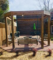 Uma pérgola de madeira com lareira e bancos, cercada por um muro de madeira e mobiliário de exterior.
