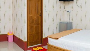 A bedroom with a wooden door, a bed with a white mattress, a TV mounted on the wall, and a small pink trash bin. - Aung Tha Pyay Hotel 2 (Yangon)
