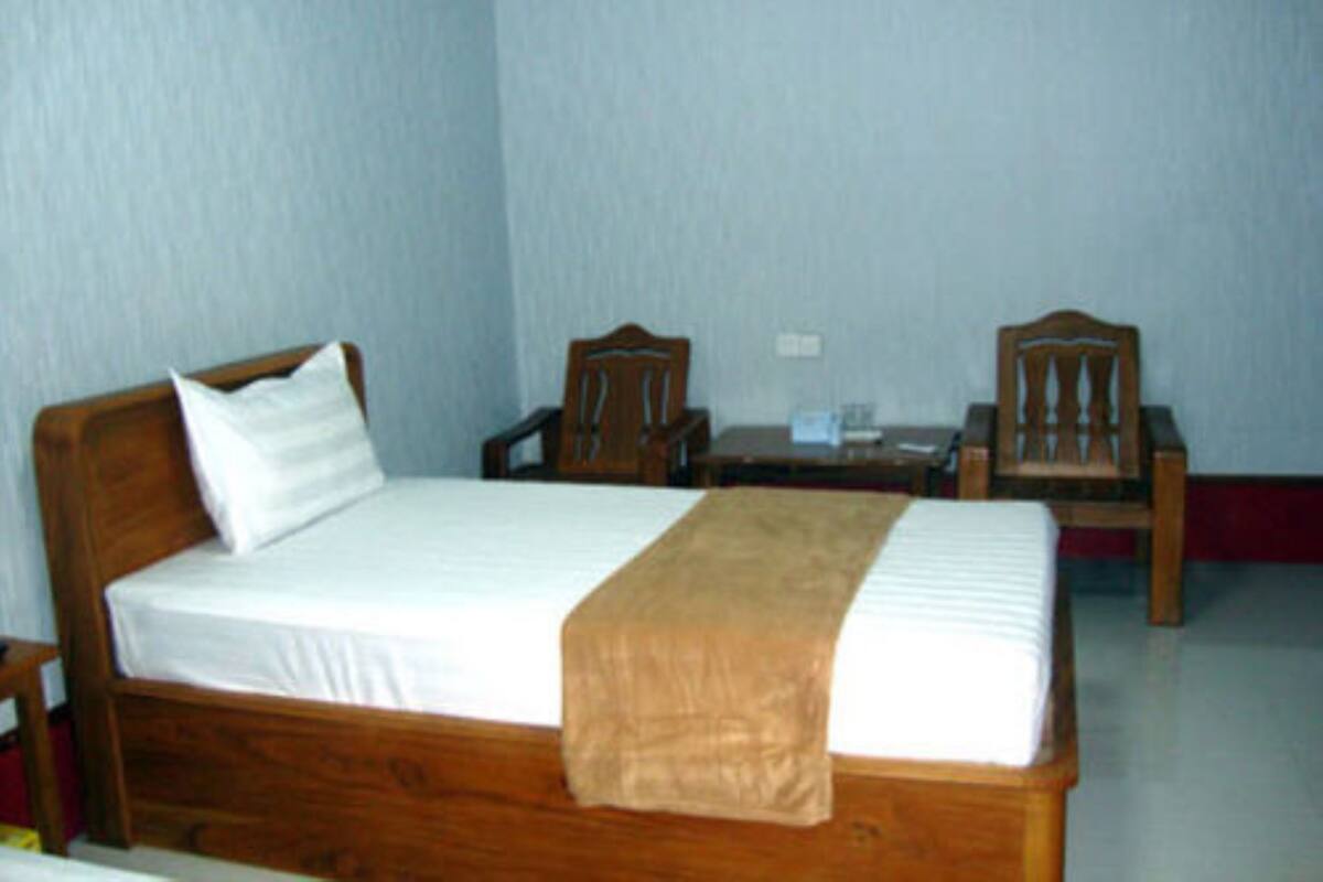 A single bed with a wooden headboard and footboard, a white mattress, and a brown throw blanket. There are two wooden chairs and a small table with a lamp.