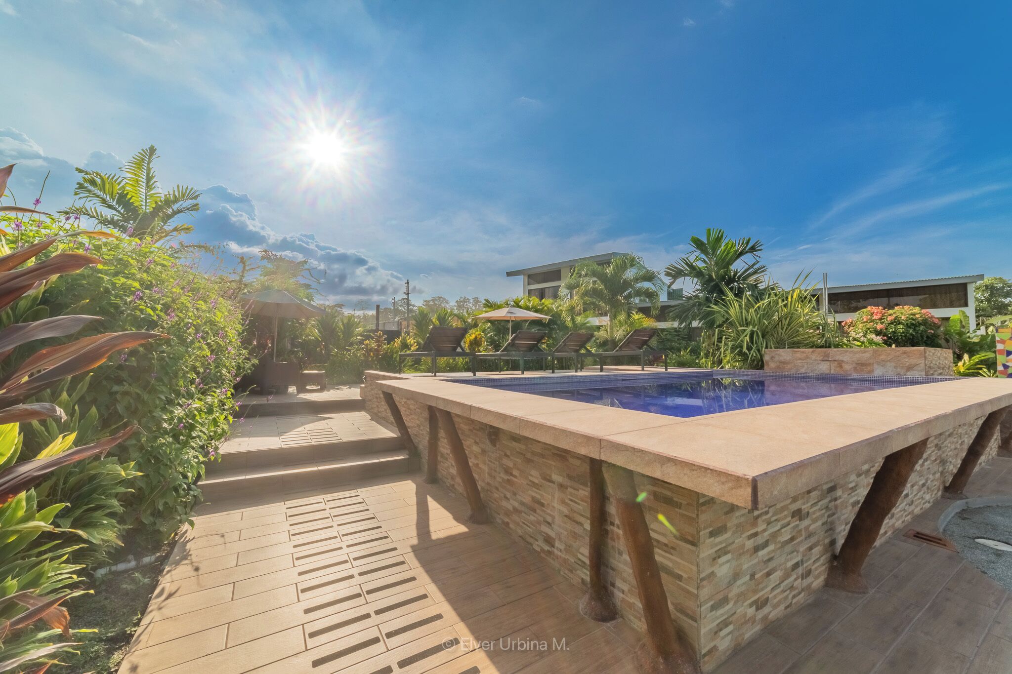 A pool area with a stone deck, surrounded by lush greenery and a modern building in the background.