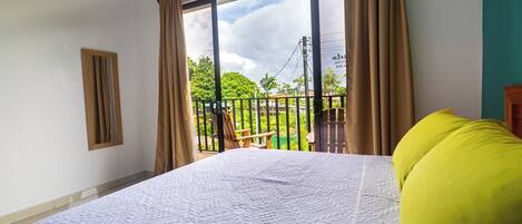 A bedroom with a large bed, a balcony with a chair, and a view of greenery.