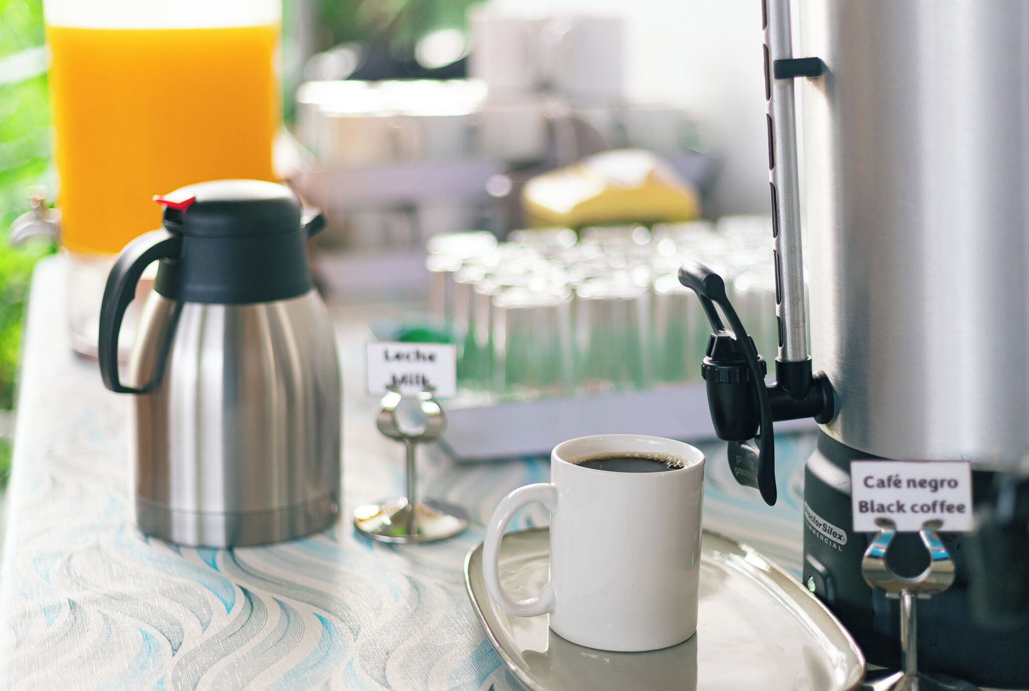 Una estación de café con una cafetera, una taza de café negro, un vaso de jugo de naranja y un cartel que dice 'Café negro Black coffee'.