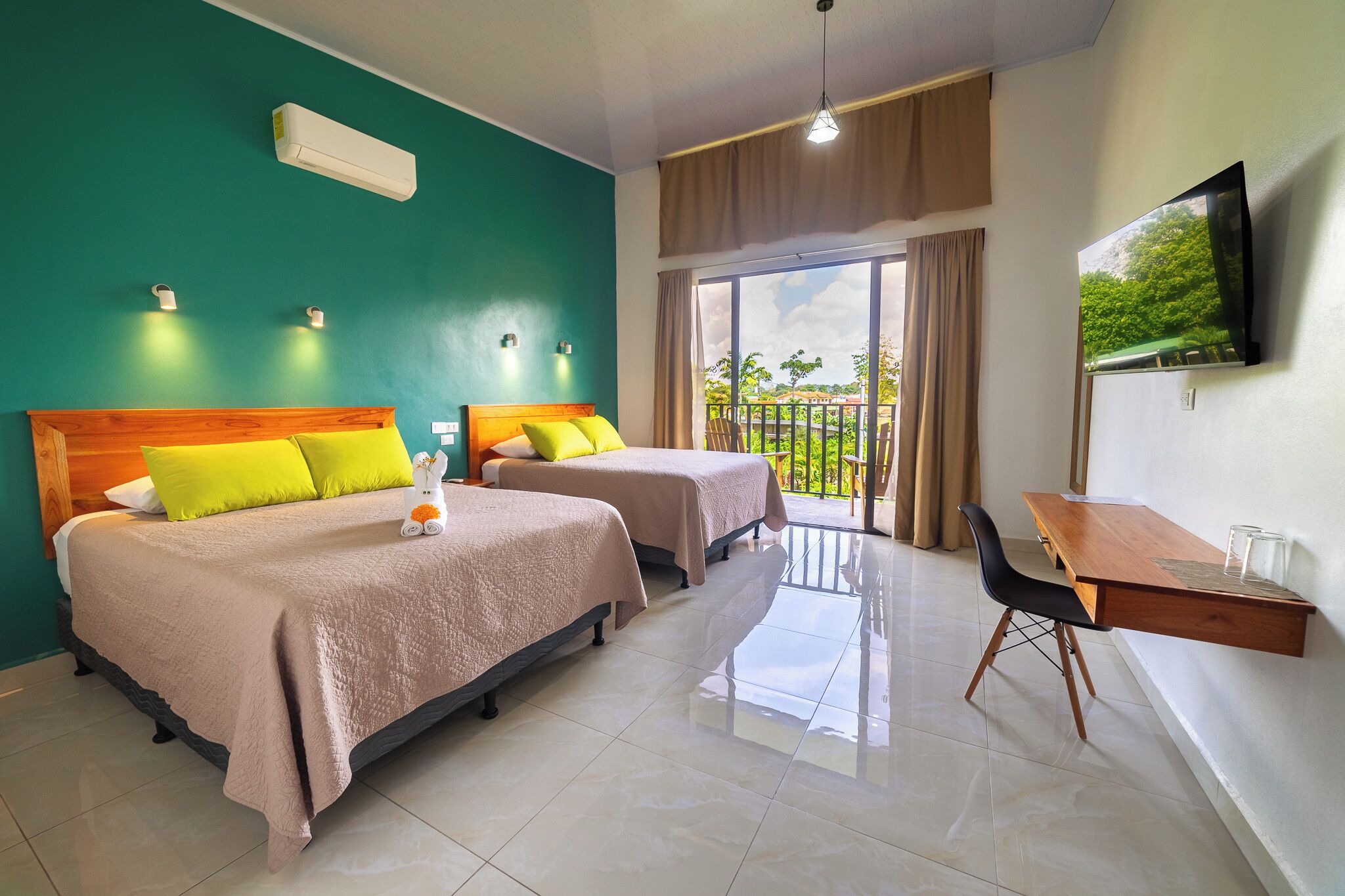 Habitación de hotel con dos camas, un escritorio, una silla, televisión y balcón con vistas a zonas verdes.