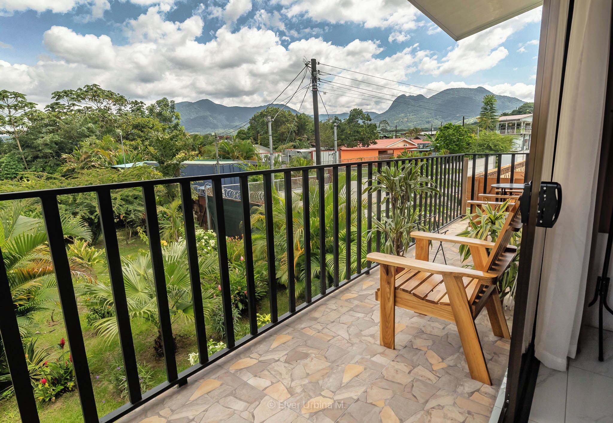 A balcony with a stone floor, wooden furniture, and a view of a lush garden and mountains.