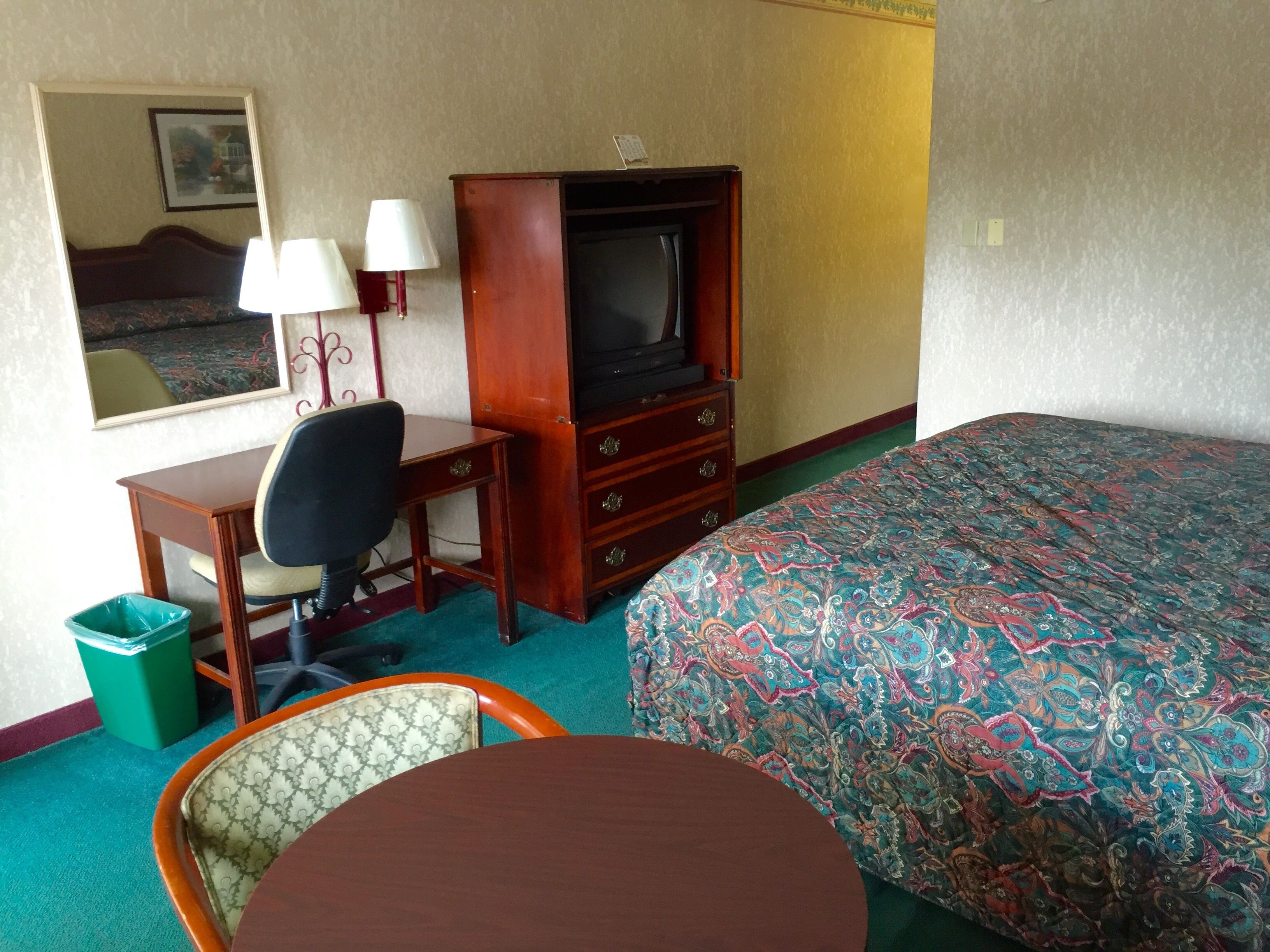 Quarto de hotel com cama, escrivaninha, cadeira, televisão e lixeira.