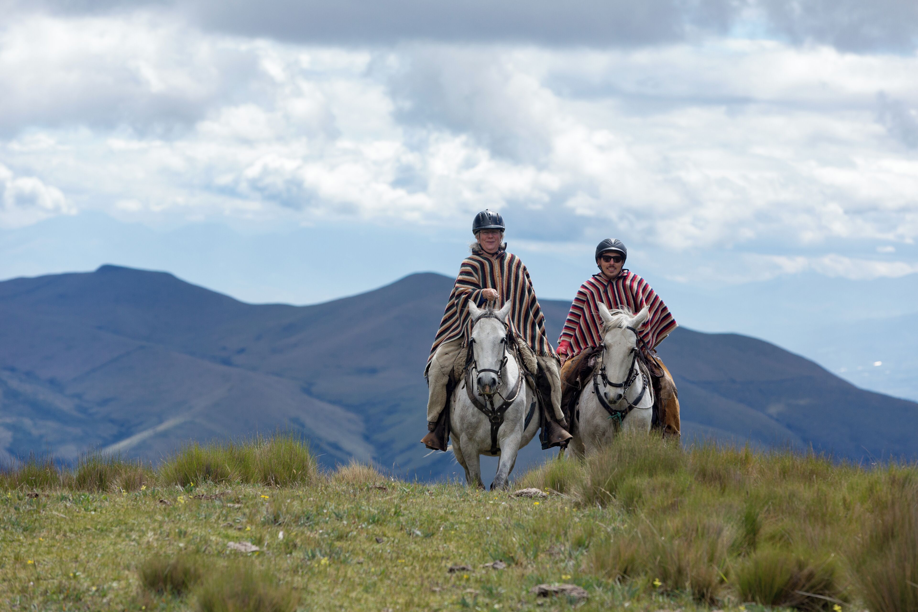 Zwei Personen reiten auf Pferden über ein grasbewachsenes Feld mit Bergen im Hintergrund.