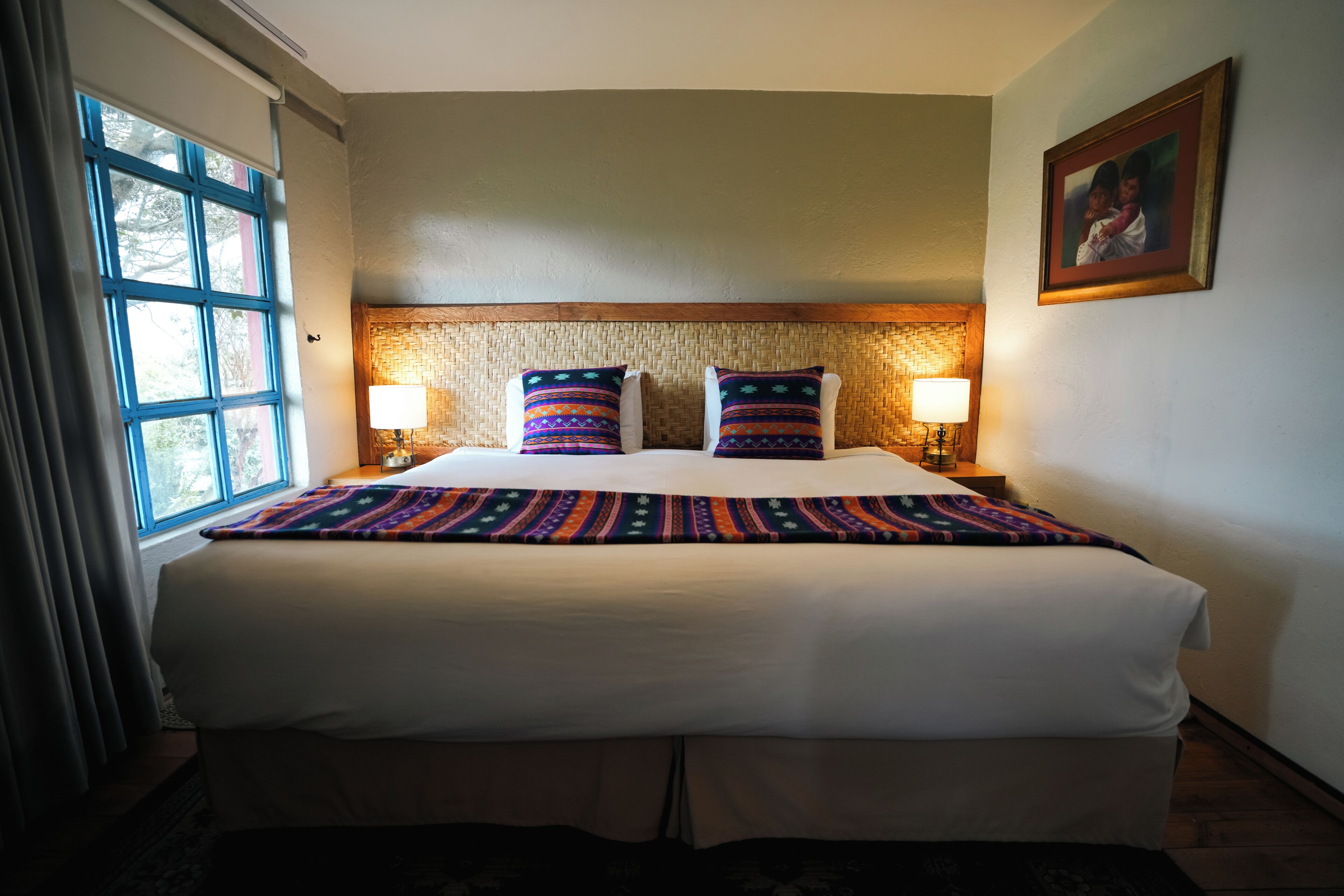 A hotel room with a large bed, two bedside lamps, a woven headboard, and a framed picture on the wall.