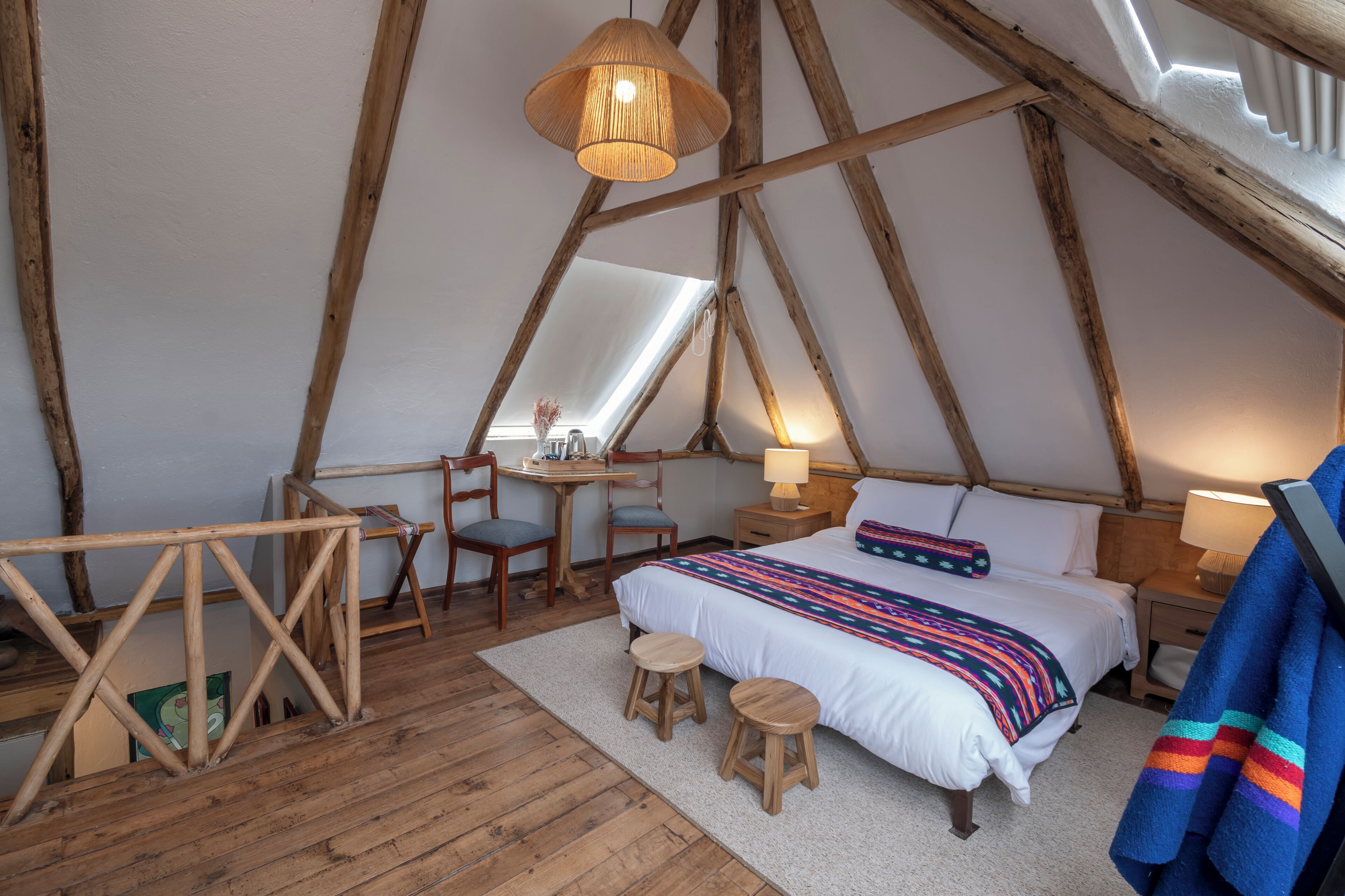 A cozy, rustic bedroom with a wooden ceiling, a bed with a colorful blanket, two stools, a desk with chairs, and a hanging light fixture.