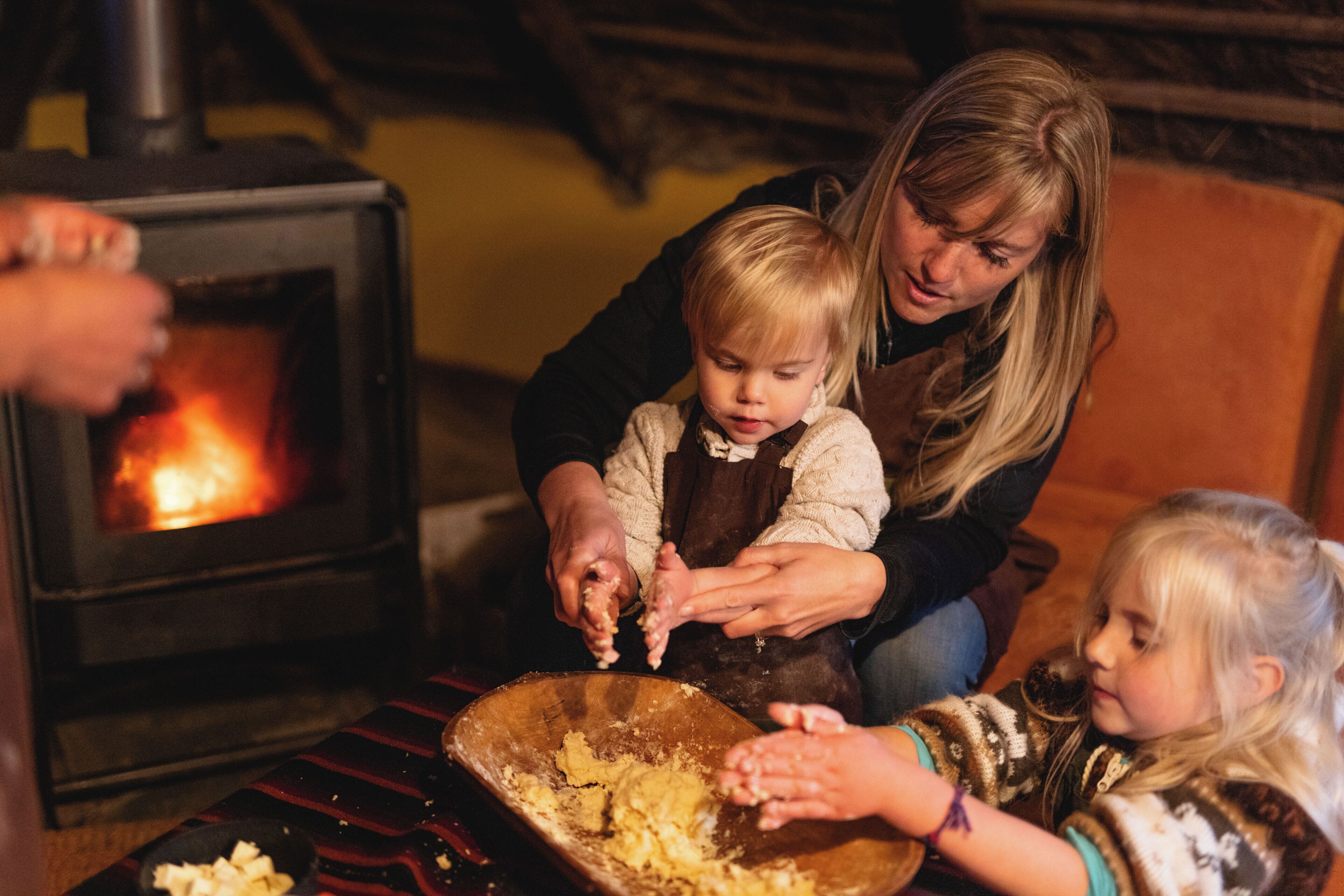 A woman and two children near a wood-burning stove, with a wooden bowl on the table.