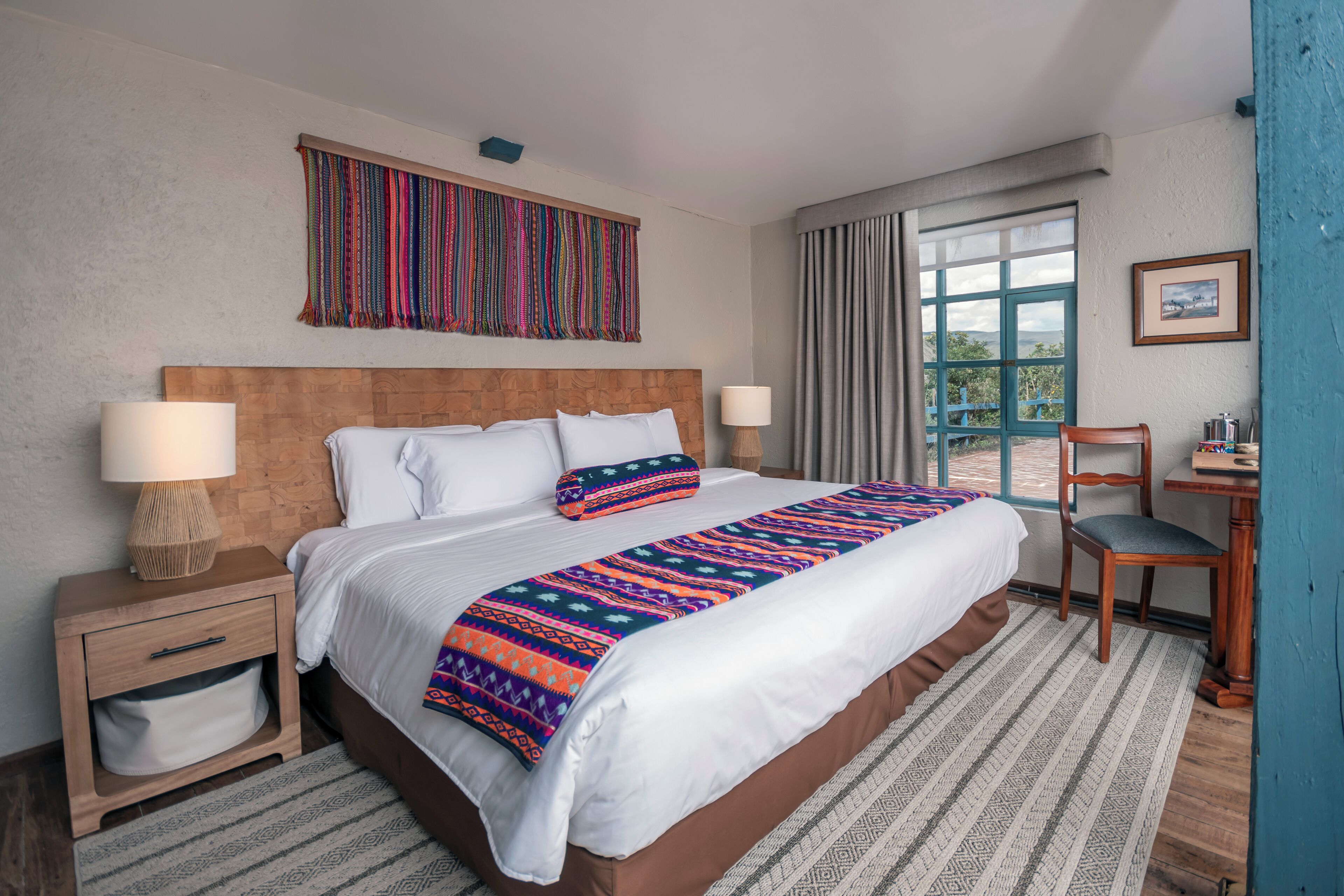 A neatly made bed with a patterned blanket, a wooden nightstand with a lamp, a chair, and a window with curtains.