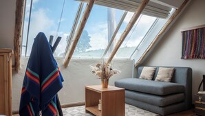 Un dormitorio estilo loft acogedor, con techo inclinado, un sofá gris, una mesita de madera con un jarrón y una manta colorida sobre una silla.