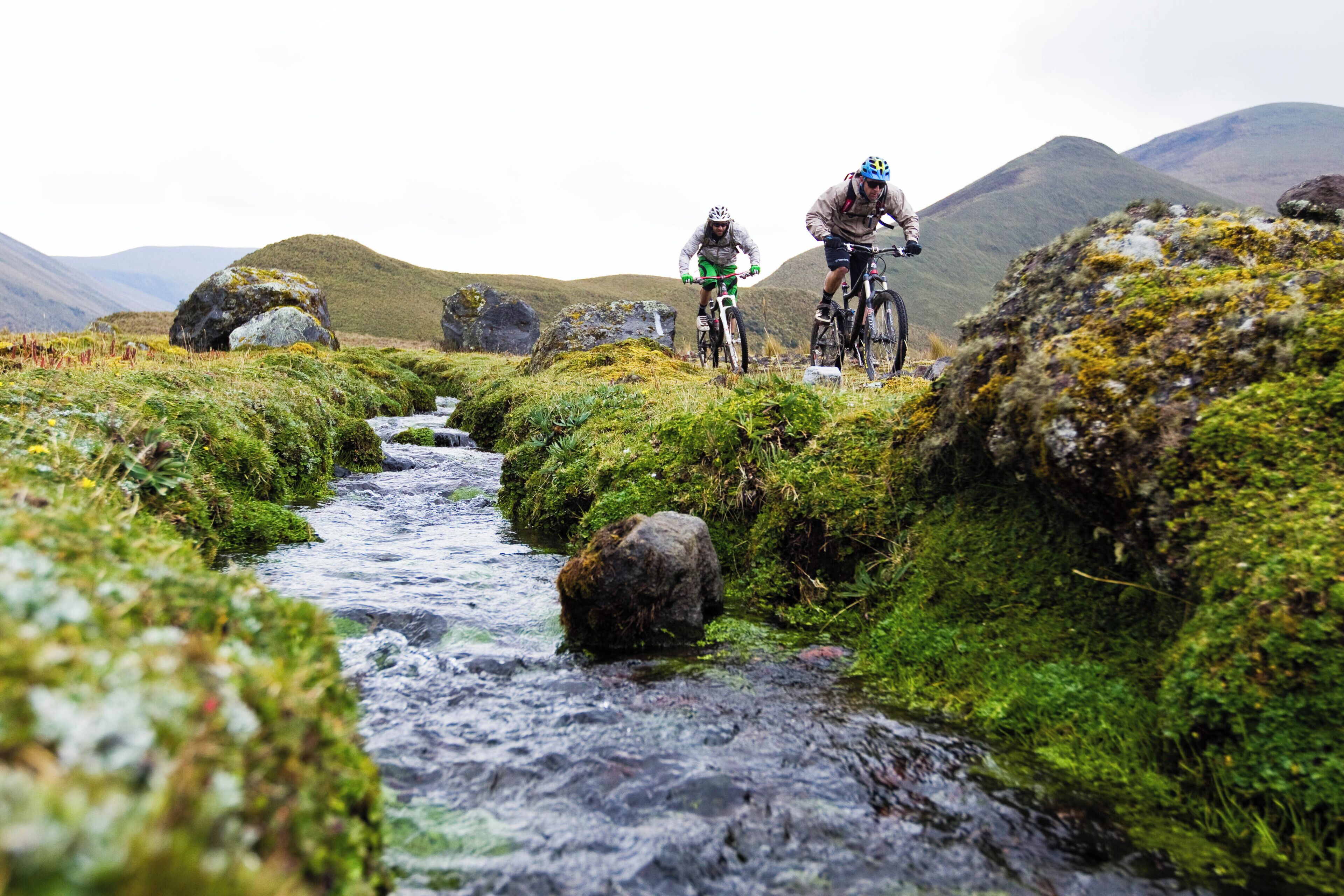 Due ciclisti da mountain bike su un sentiero con un ruscello e terreno roccioso.