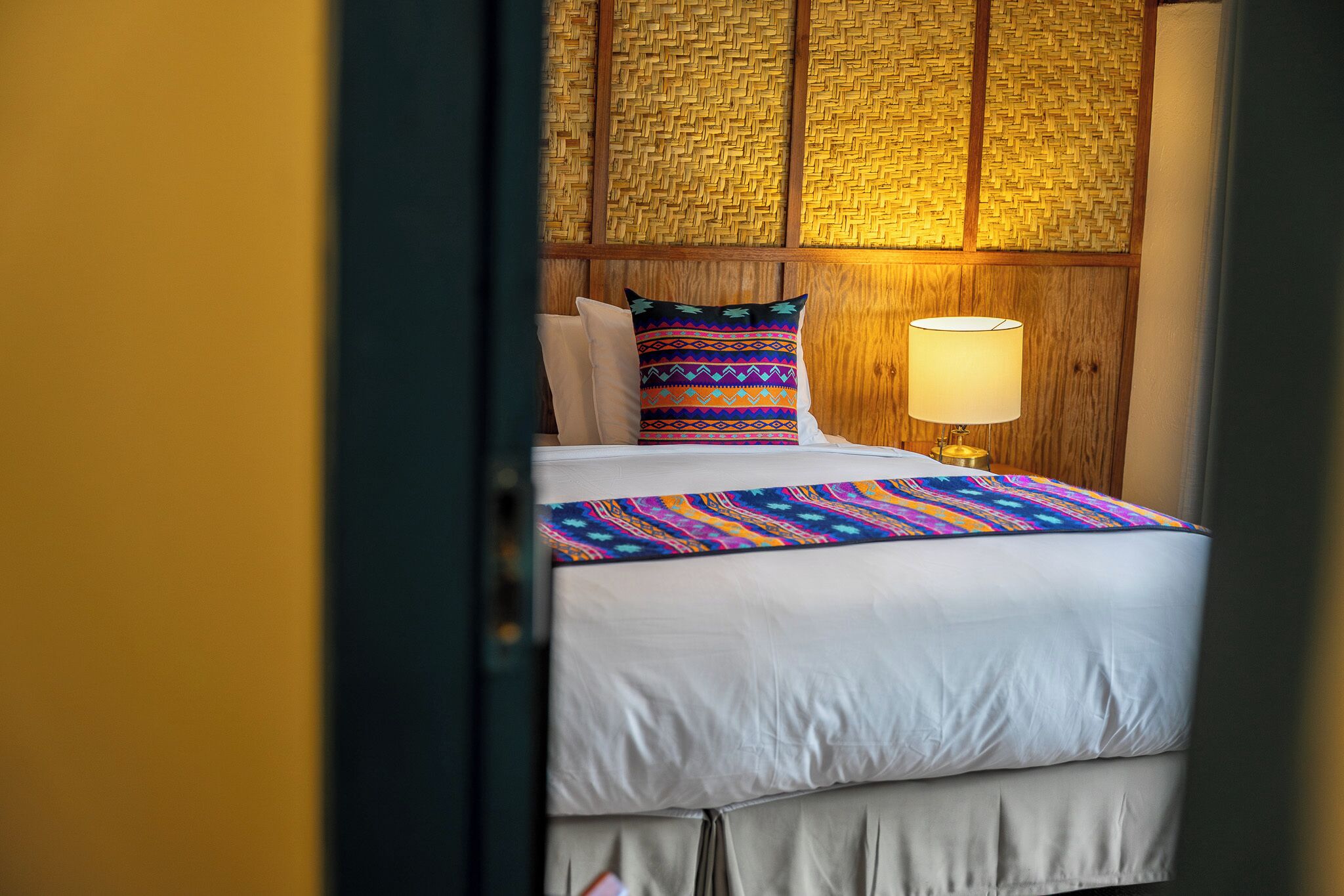 A bed with a colorful blanket and pillows, a bedside lamp, and a woven headboard.