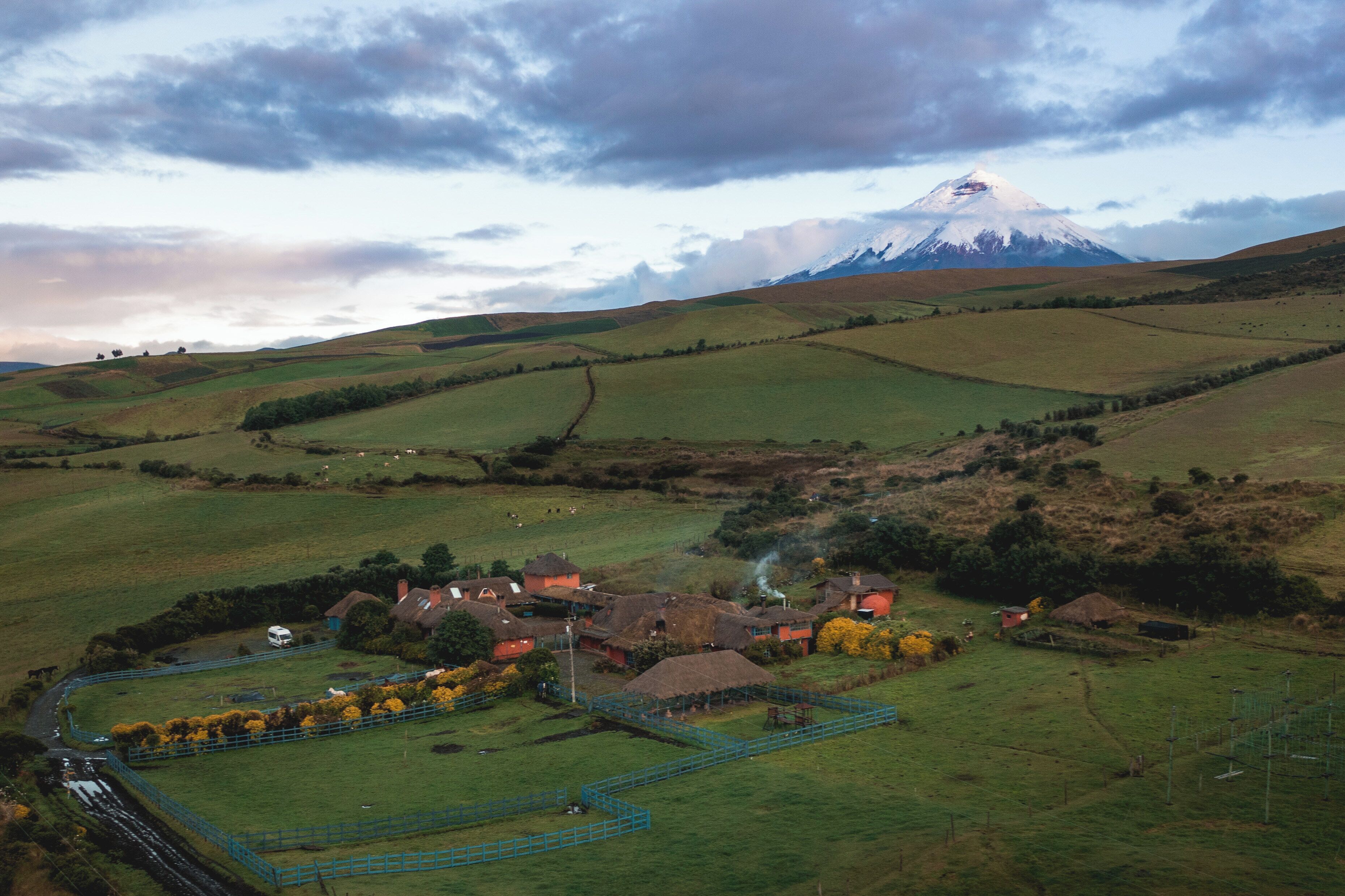 Un paysage rural avec une montagne en arrière-plan, un petit village avec des maisons et un bâtiment à toit de chaume, et une zone délimitée par une clôture où se trouvent des arbres.