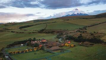Paisaje rural con una montaña al fondo, un pequeño pueblo con casas y un edificio de techo de paja, y un área cercada con árboles.