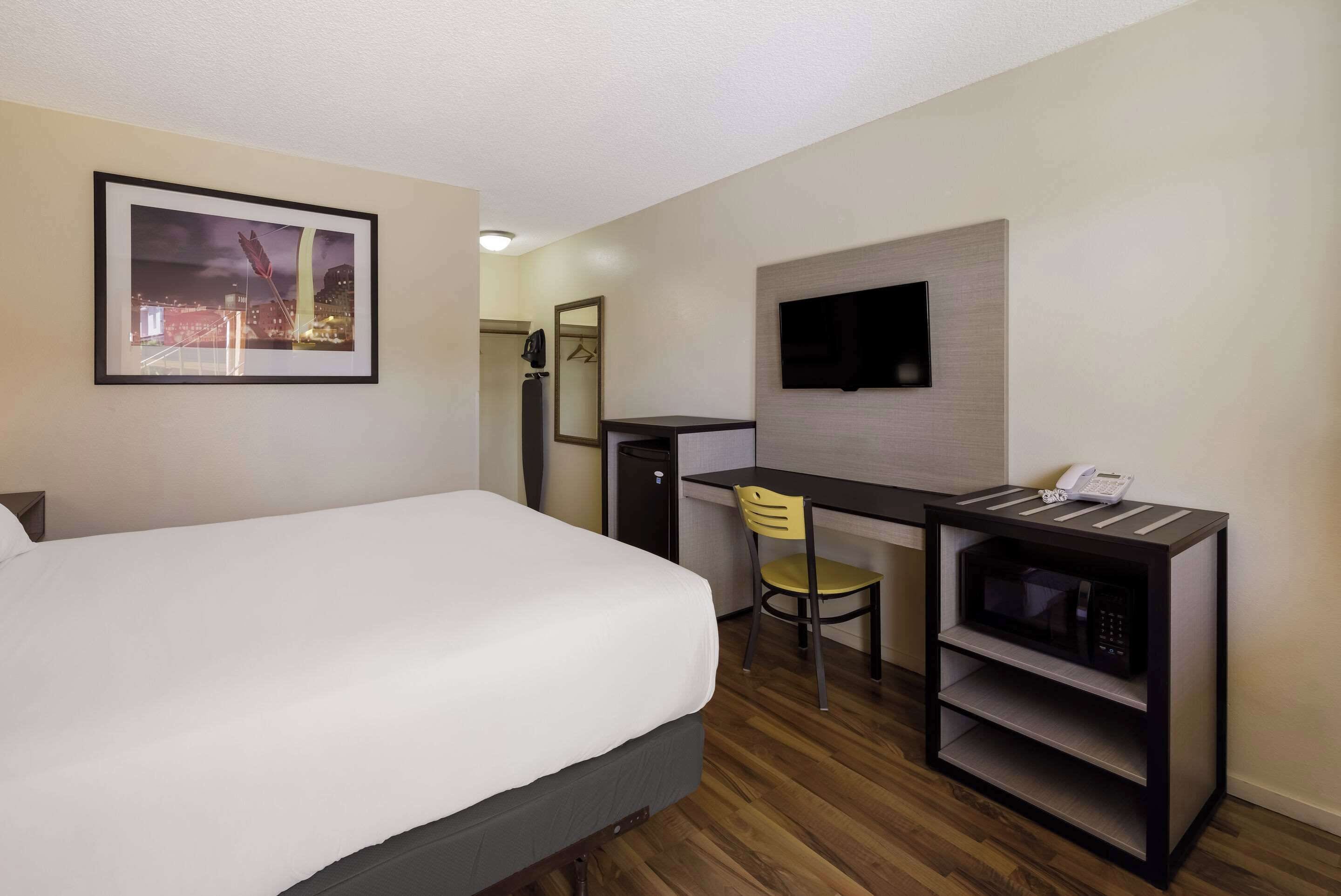 Habitación de hotel con cama, escritorio con silla, microondas, televisión y un cuadro en la pared.