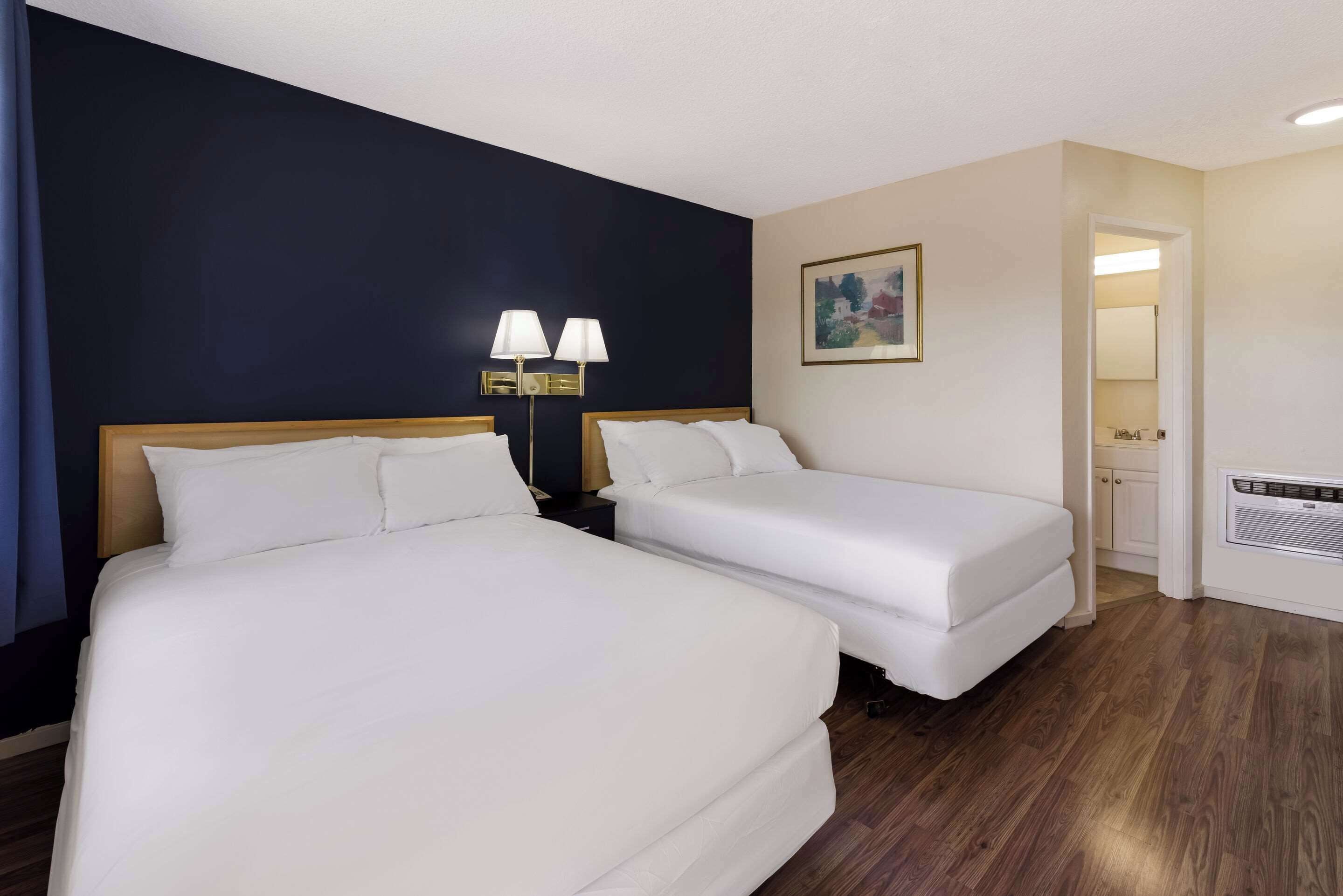 A hotel room with two beds, a dark accent wall, a painting, and a bathroom door.