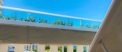 A balcony with a white chair, a small table, and a view of a clear blue sky and greenery.
