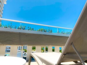 A balcony with a white chair, a small table, and a view of a clear blue sky and greenery. - The Wave Hotel Condado (San Juan)