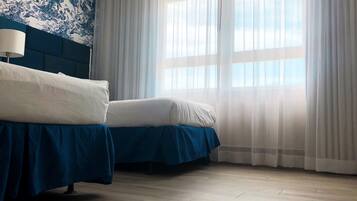 A hotel room with two beds, a blue headboard, a large window with sheer curtains, and a patterned wall.