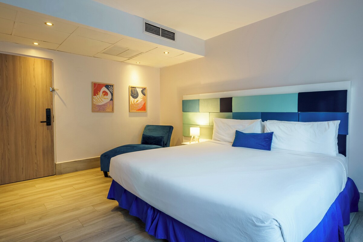 A hotel room with a large bed, a chair, a door, and two wall-mounted artworks.