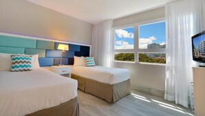 Habitación de hotel con dos camas, una mesita de noche, una lámpara, un televisor y un ventanal con vistas a edificios y árboles.