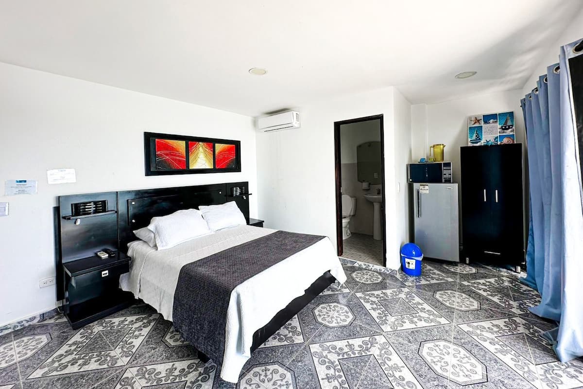 A hotel room with a bed, a small desk, a wardrobe, a bathroom visible through an open door, and patterned floor tiles.