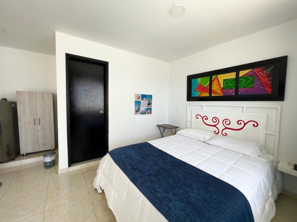 Ein Schlafzimmer mit einem Bett, einer Tür, einem kleinen Tisch, einem Schrank und einer Wand mit einem farbenfrohen abstrakten Gemälde.