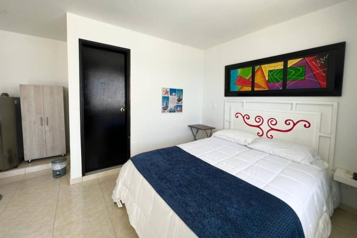 A bedroom with a bed, a door, a small table, a cabinet, and a wall with a colorful abstract painting.