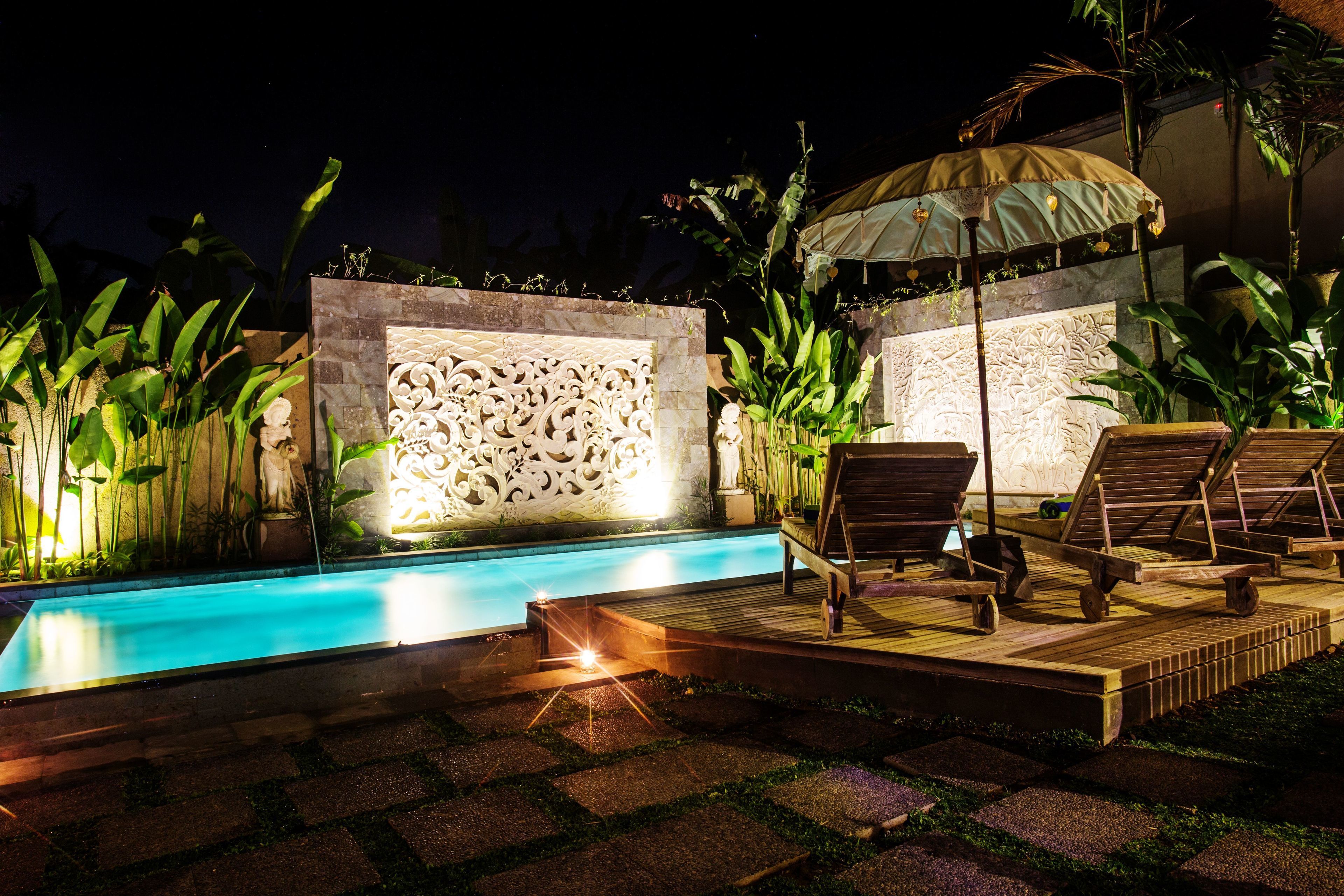 A nighttime pool area with a decorative wall, lounge chairs, and an umbrella.