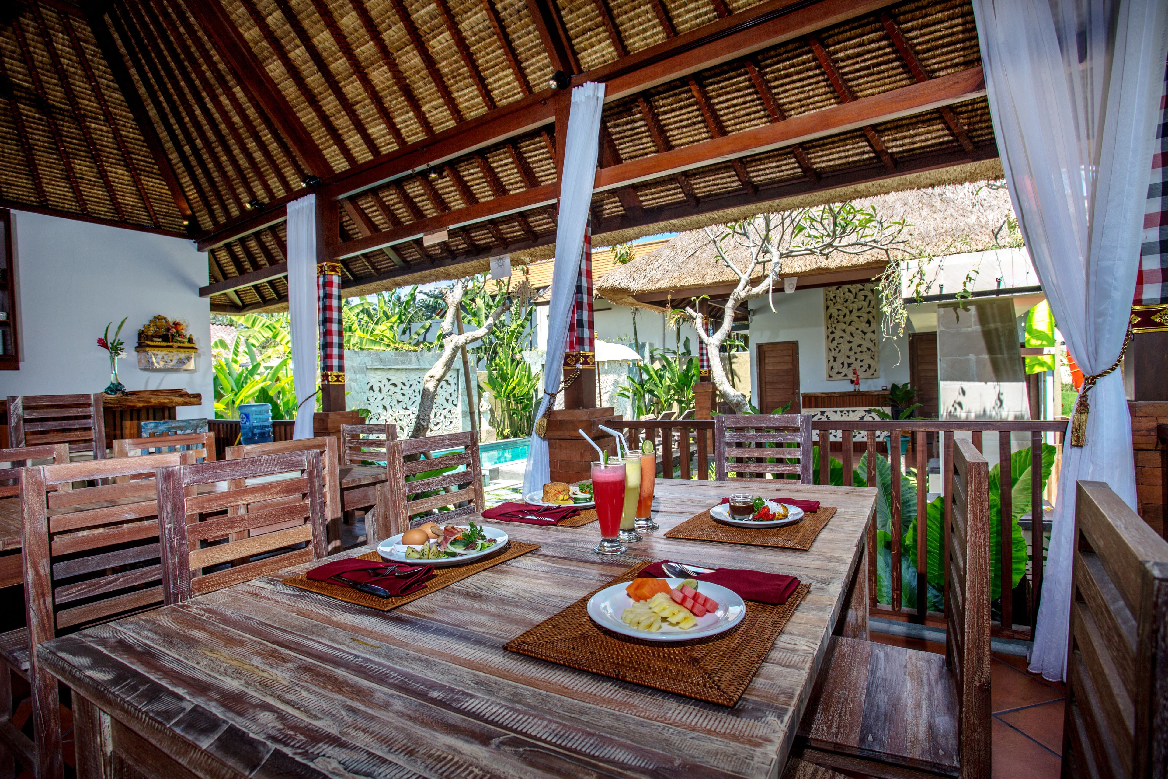 An outdoor dining area with wooden tables and chairs, surrounded by lush greenery and a thatched roof structure.