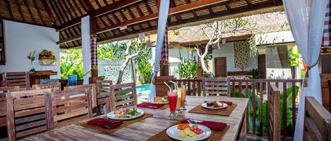 An outdoor dining area with wooden tables and chairs, surrounded by lush greenery and a thatched roof structure.