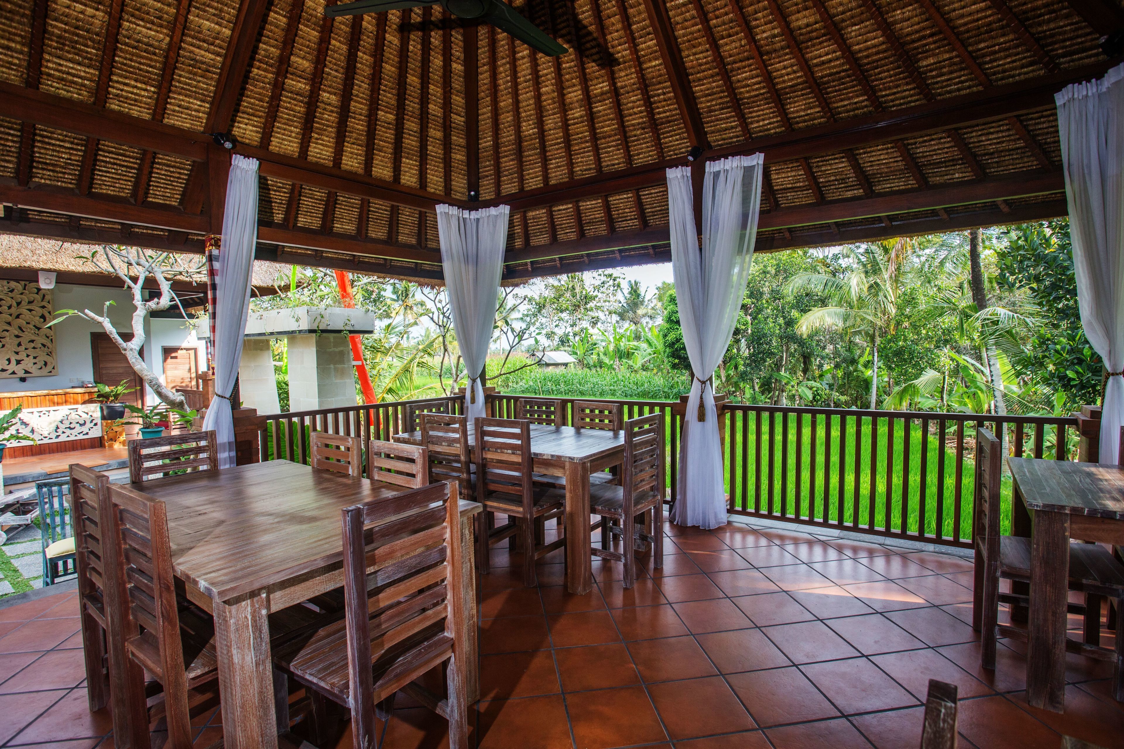 An outdoor patio with wooden tables and chairs, a thatched roof, and a view of lush greenery.