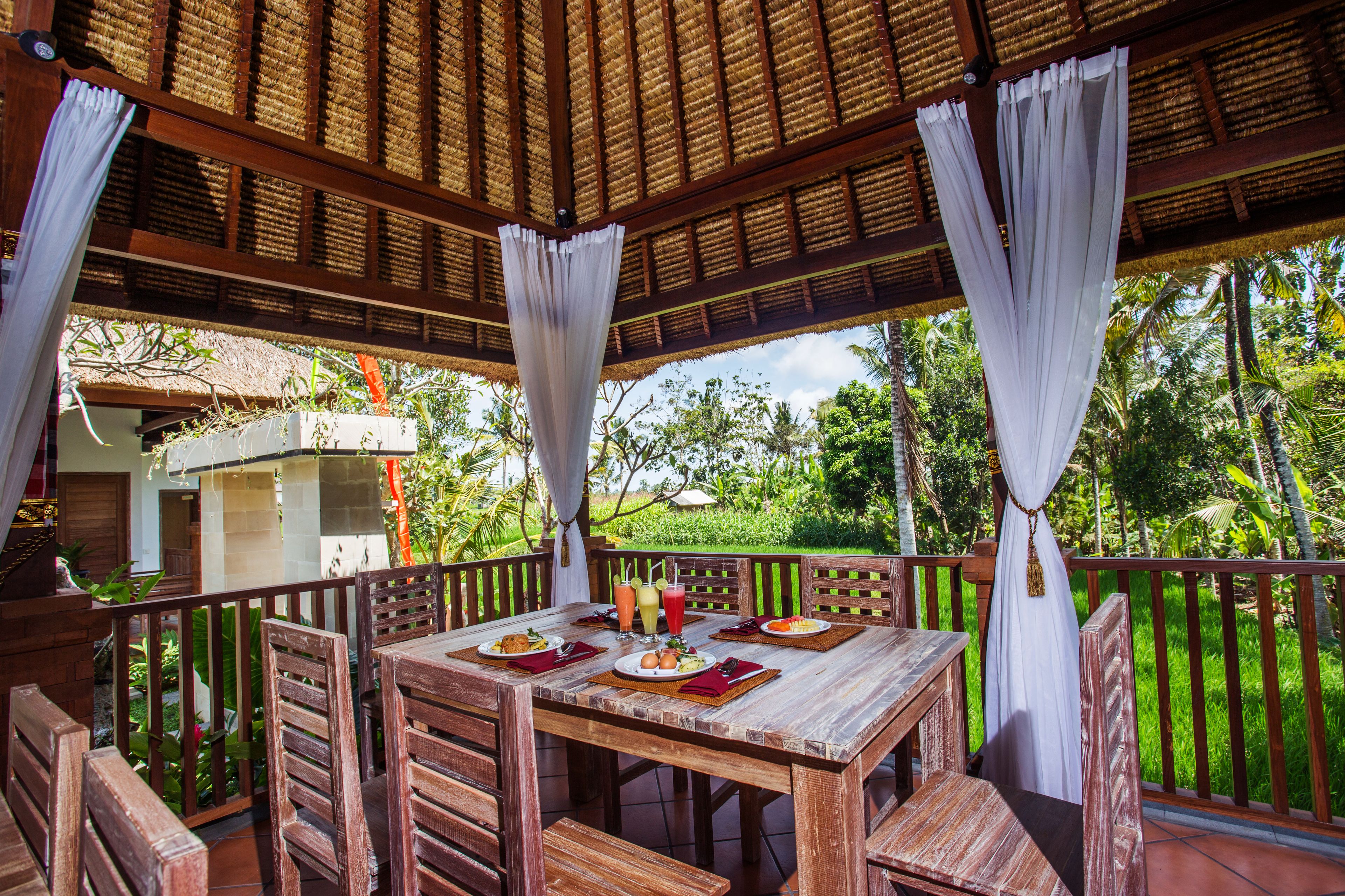 A wooden outdoor dining area with a table set for a meal, surrounded by chairs, and a thatched roof overhead.