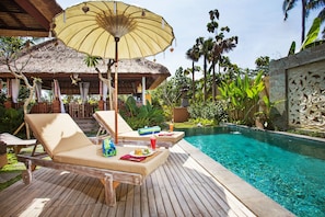 Une terrasse en bois avec des chaises longues et un parasol, une piscine et une structure à toit de chaume.