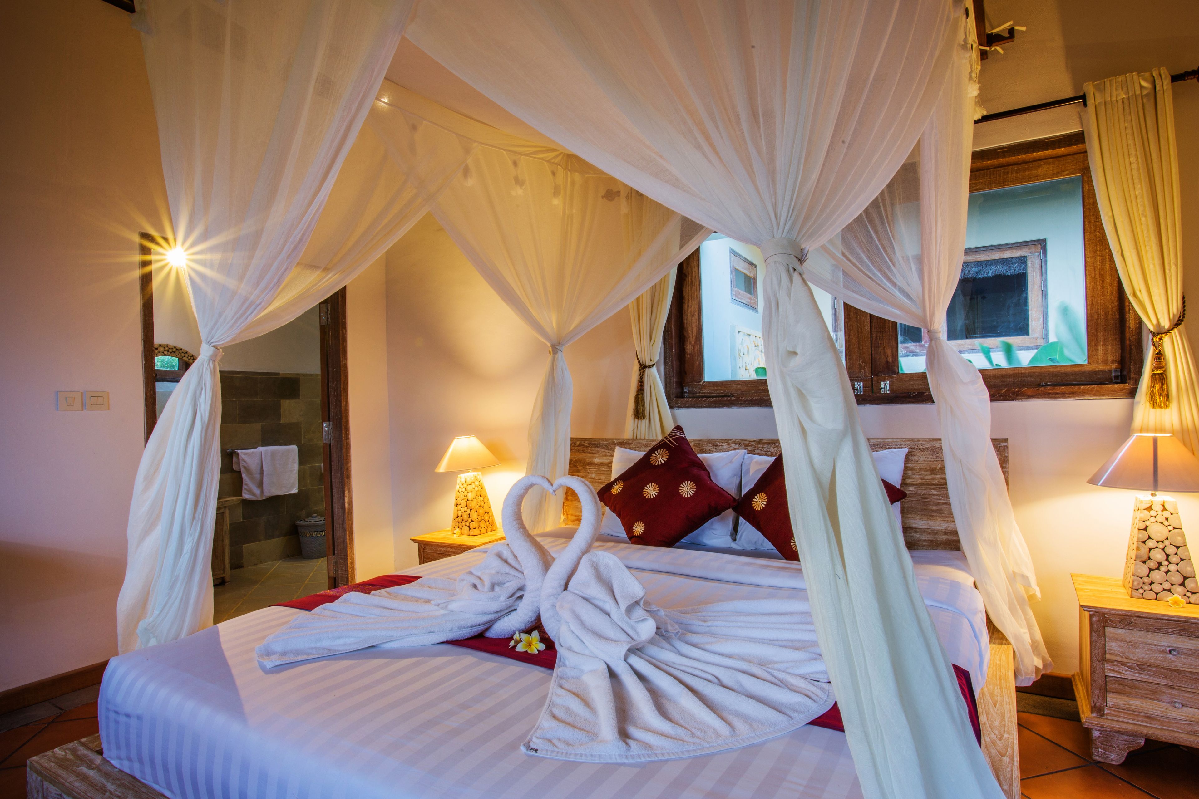 A bed with a canopy, towels arranged in a heart shape, and a nightstand with a lamp.