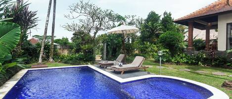 Une piscine avec deux chaises longues et un parasol, le tout entouré d’une végétation luxuriante, avec une maison en arrière-plan.
