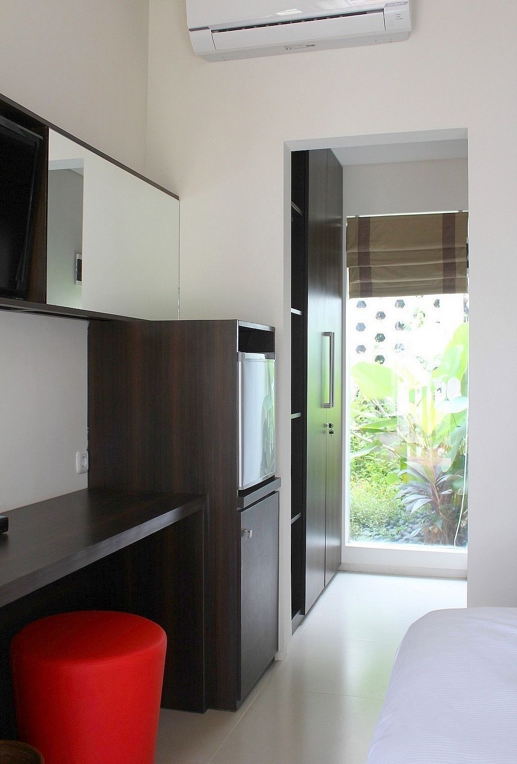 A compact hotel room with a kitchenette, a bed, and a view of greenery through a glass door.
