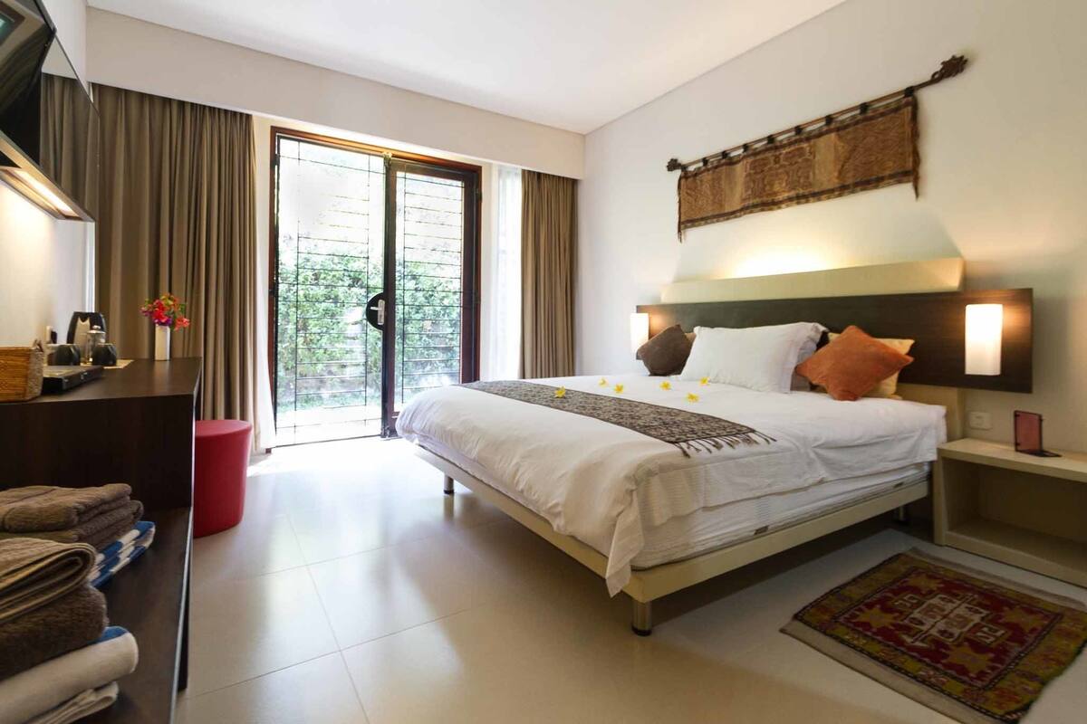 A hotel room with a bed, a desk, a chair, a red trash bin, and a view of the outdoors through a sliding door.