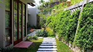 A narrow pathway leading to a building with large glass doors, surrounded by lush greenery and a stone wall. - Villa Karingal (Senggigi)