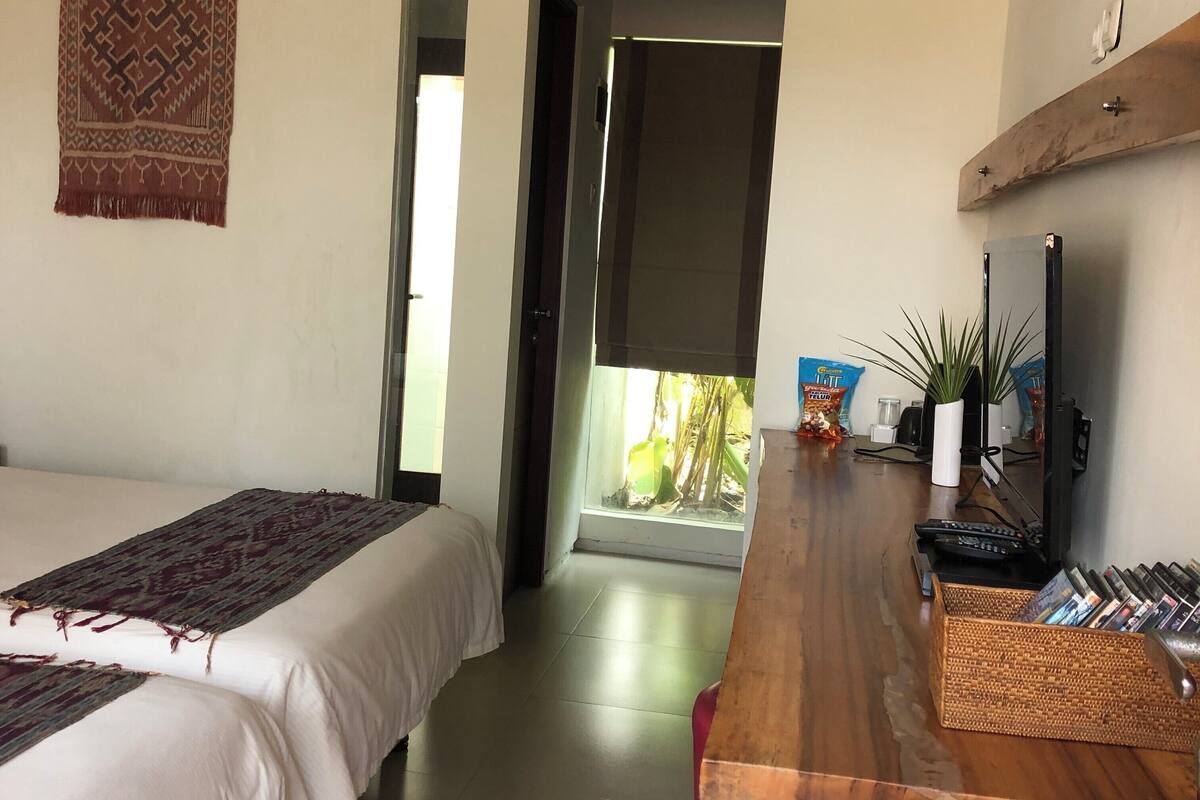 A hotel room with two beds, a wooden desk with a computer and a plant, and a view of greenery outside.
