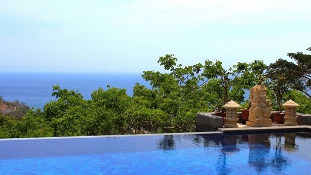 An infinity pool with a view of the ocean, surrounded by lush greenery and decorative stone statues. - Villa Karingal (Senggigi)