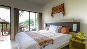 A hotel room with a large bed, a nightstand with a vase of flowers, and a view of a garden and buildings outside. - Villa Karingal (Senggigi)