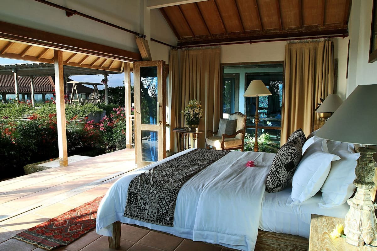 A bedroom with a large bed, a chair, a table, and a view of the outdoors.