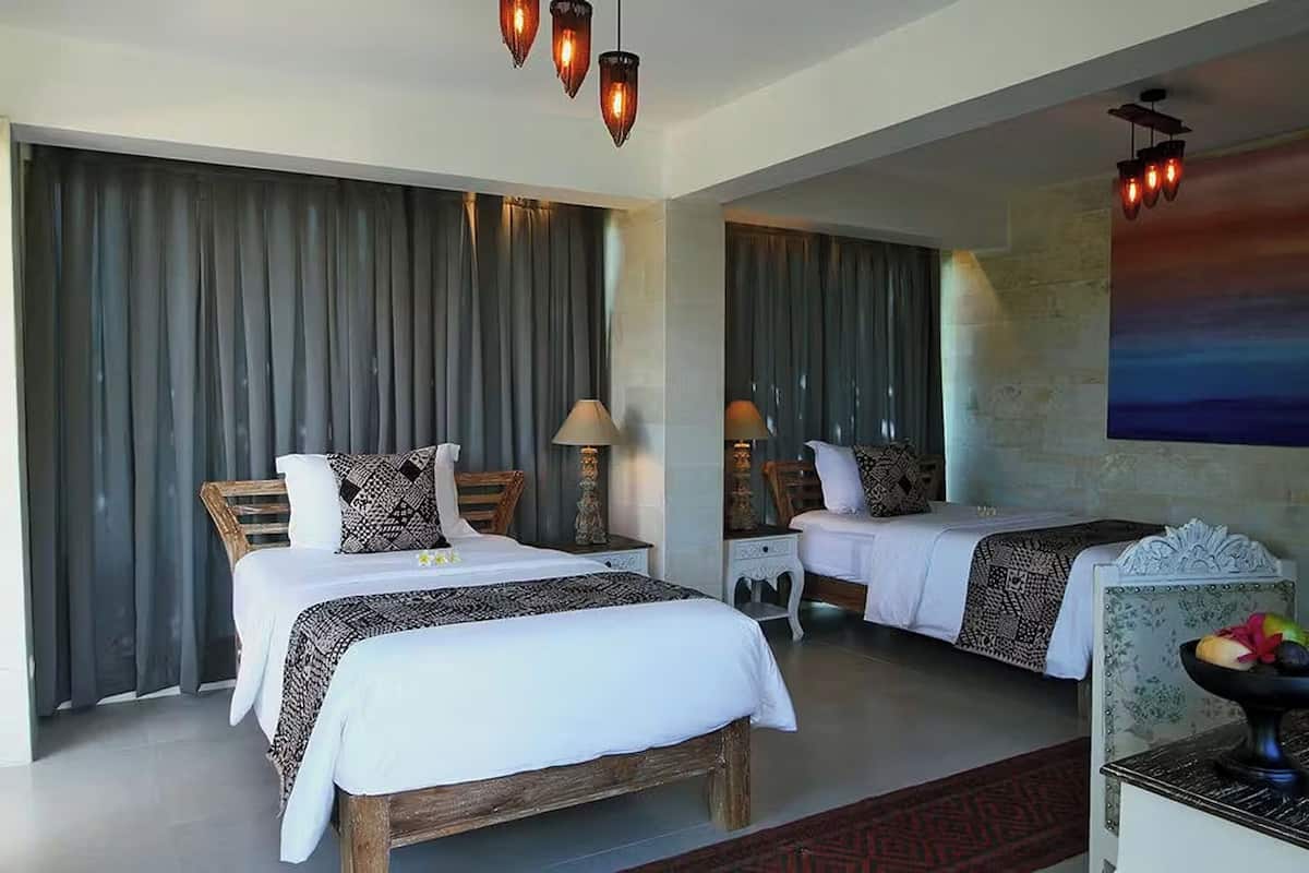 A hotel room with two beds, a large window with curtains, a painting on the wall, and a small table with a bowl of fruit.