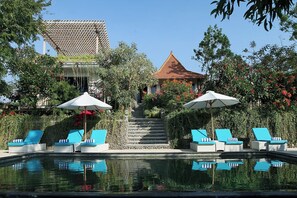 A pool area with lounge chairs and umbrellas, surrounded by lush greenery and a traditional building with a tiled roof. - Bangsring Breeze (Banyuwangi)